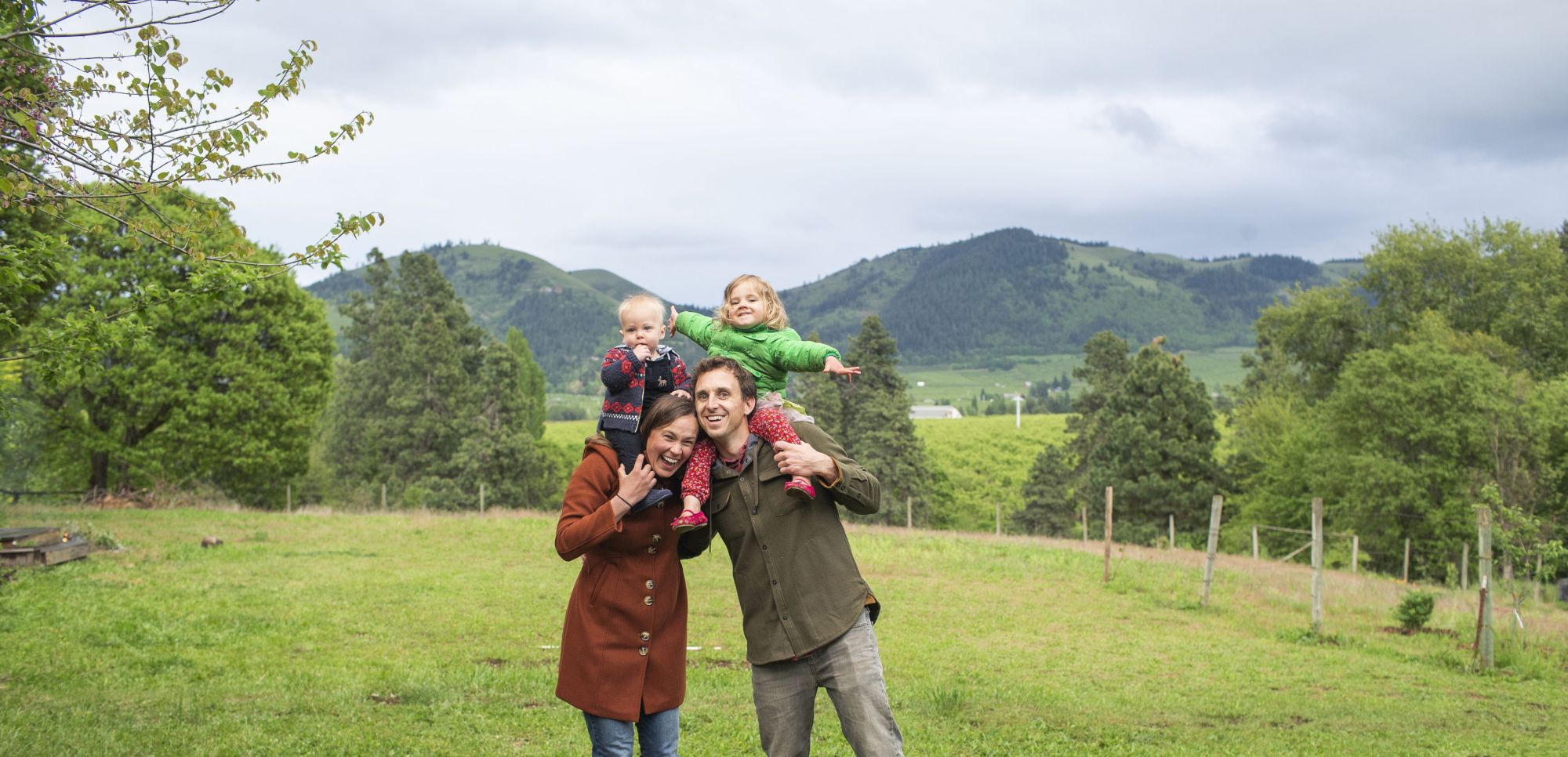A family of four is posing in a grassy field with mountains in the background. The children are on the adults' shoulders.