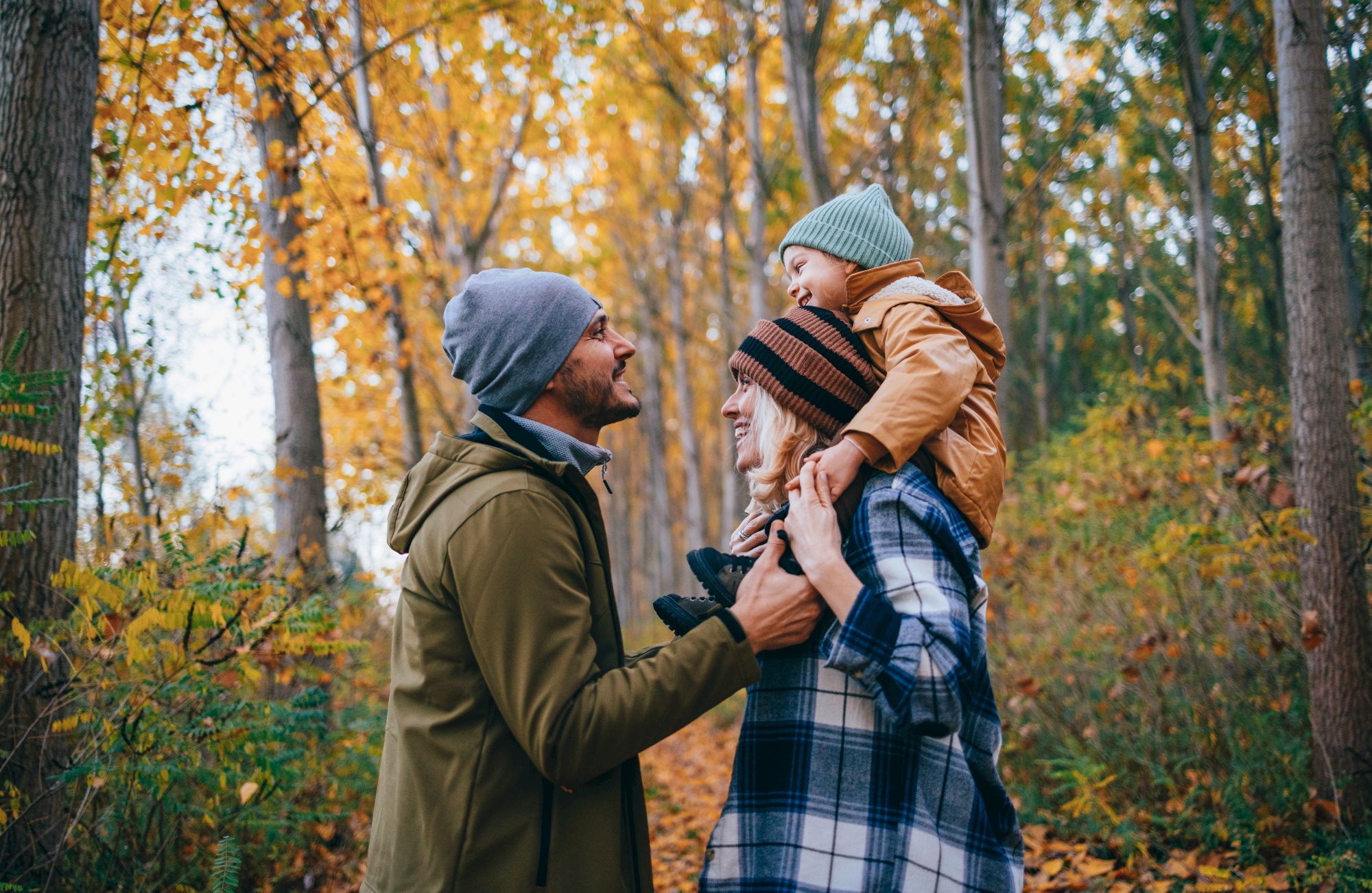A family in autumn attire shares a moment in a forest, surrounded by trees with golden leaves, enjoying a walk together.