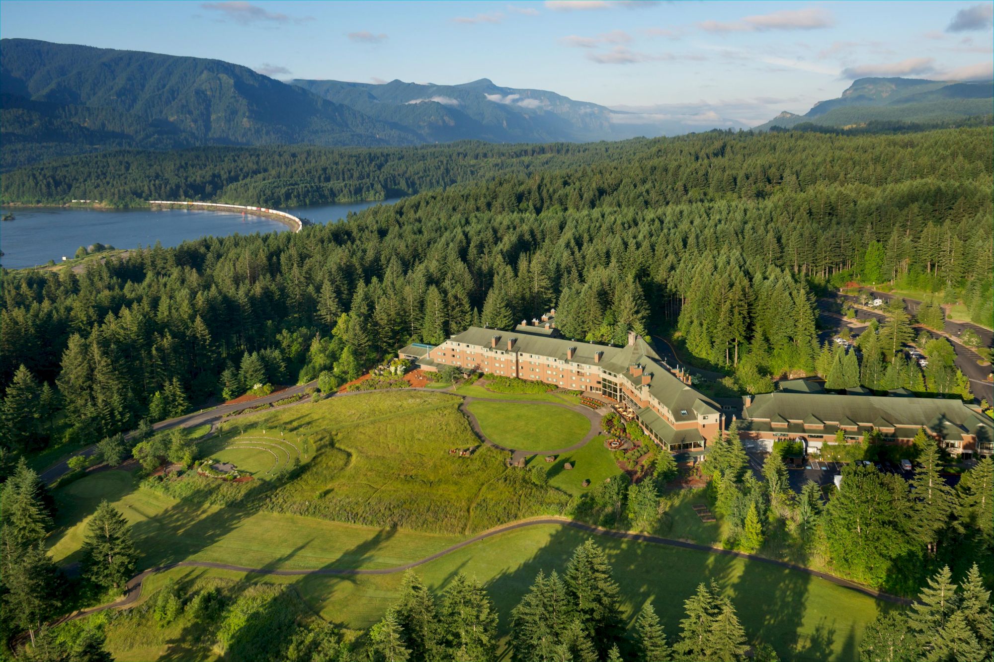 An expansive aerial view of a large building surrounded by lush forests, with a river and mountain scenery in the background.