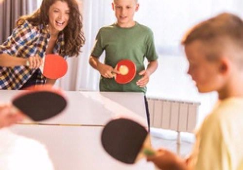 People are playing a doubles ping pong game indoors, appearing engaged and having fun with bright lighting in the background.