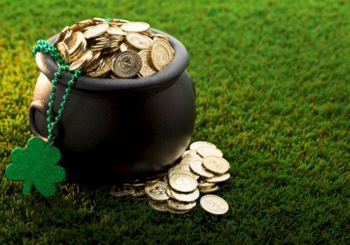 A pot filled with gold coins, a shamrock necklace hangs on the side, placed on lush green grass.
