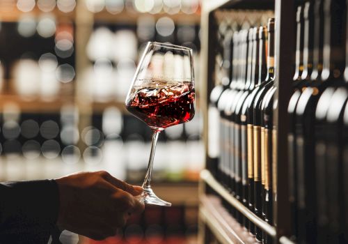 A hand holds a glass of red wine in a wine store, with shelves of wine bottles in the background, creating a sophisticated ambiance.
