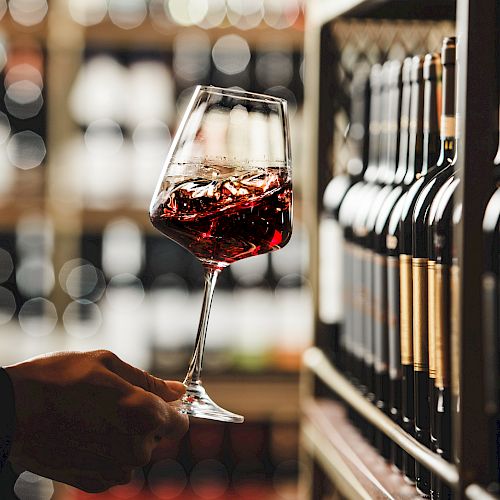 A hand holds a glass of red wine in a wine store, with shelves of wine bottles in the background, creating a sophisticated ambiance.