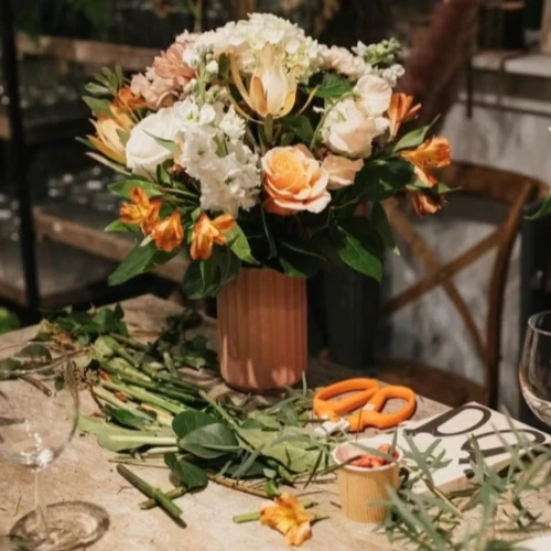 A flower arrangement with lilies and roses is on a table, with scissors and wine glasses nearby, set in what seems like a cozy space.