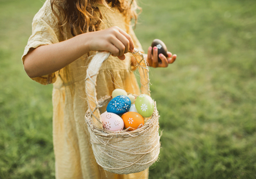 A person in a yellow dress holds a basket filled with colorful decorated eggs, standing on lush green grass, signifying an Easter egg hunt.