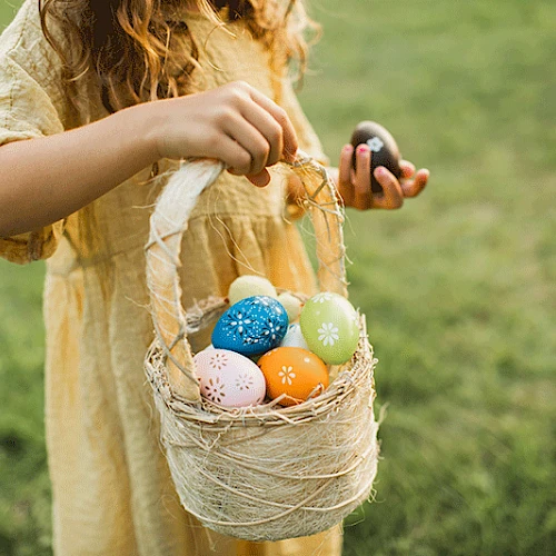 A person in a yellow dress holds a basket filled with colorful decorated eggs, standing on lush green grass, signifying an Easter egg hunt.