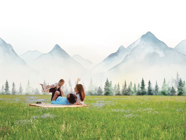 People having a picnic on a blanket in a grassy field, with a forest and distant mountains in the background.