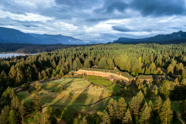An aerial view of a large building surrounded by a forest, with mountains in the distance and a body of water to the left.