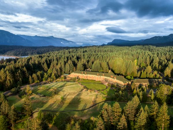 An aerial view of a large building surrounded by a forest, with mountains in the distance and a body of water to the left.