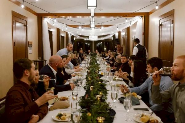 The image shows a long dining table with many people enjoying a meal in a decorated room with string lights and greenery.