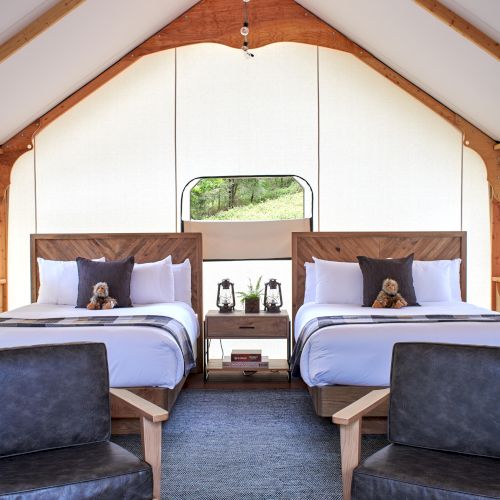 The image shows a cozy room with two beds, chairs, and a small table with board games in a brightly lit, tent-like structure.