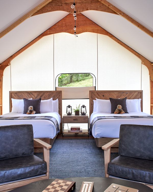 The image shows a cozy room with two beds, chairs, and a small table with board games in a brightly lit, tent-like structure.