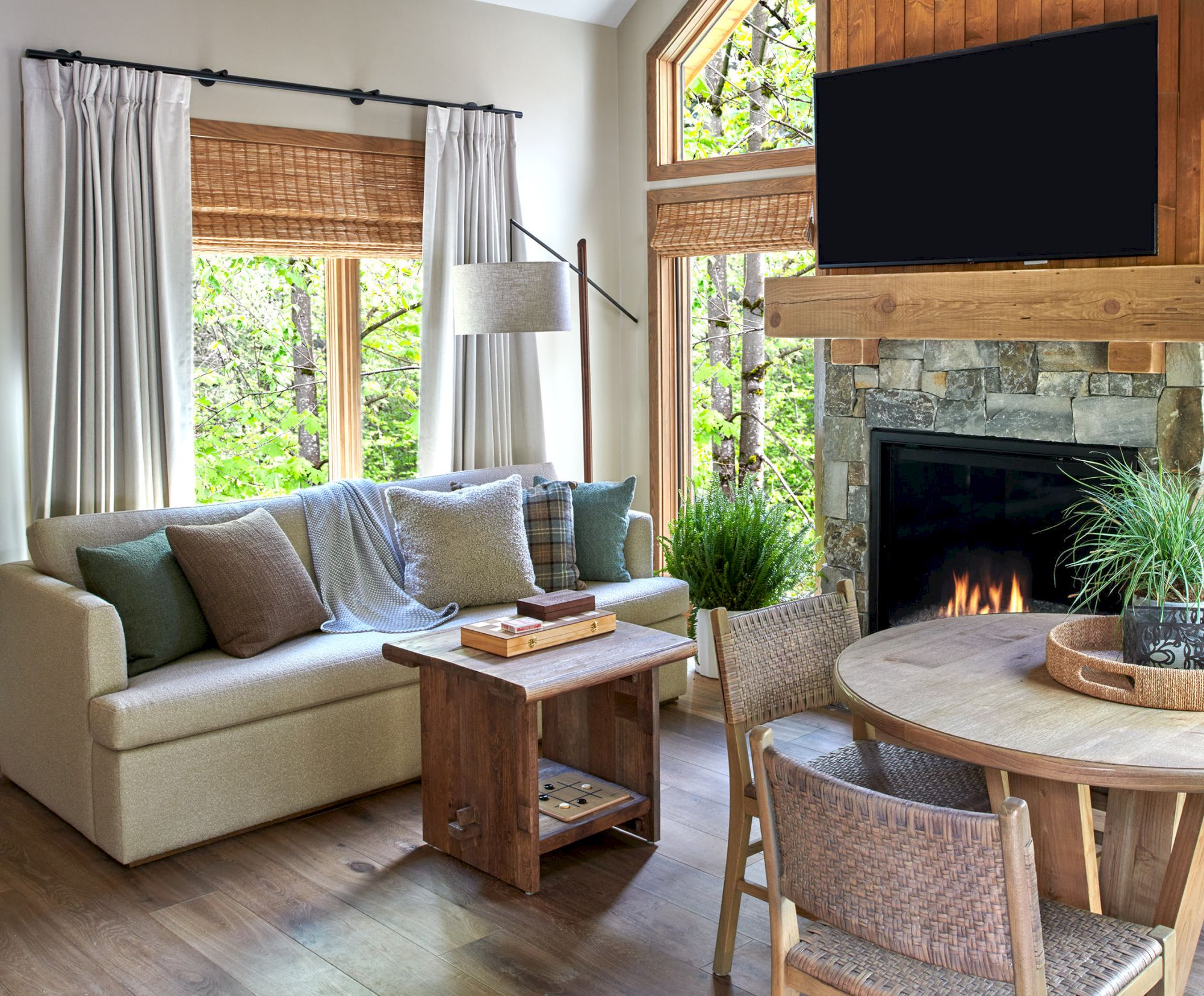 A cozy living room with a beige sofa, pillows, a wooden table, fireplace, TV, round dining table, and large windows with greenery outside.