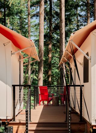 Two white tents with orange roofs face each other, separated by a wooden deck with a red chair, surrounded by tall trees in the background.