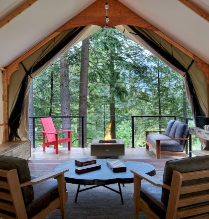 The image shows a cozy cabin interior with a view of trees. It has wooden chairs, a fire pit, couches, and an open front creating a serene setting.