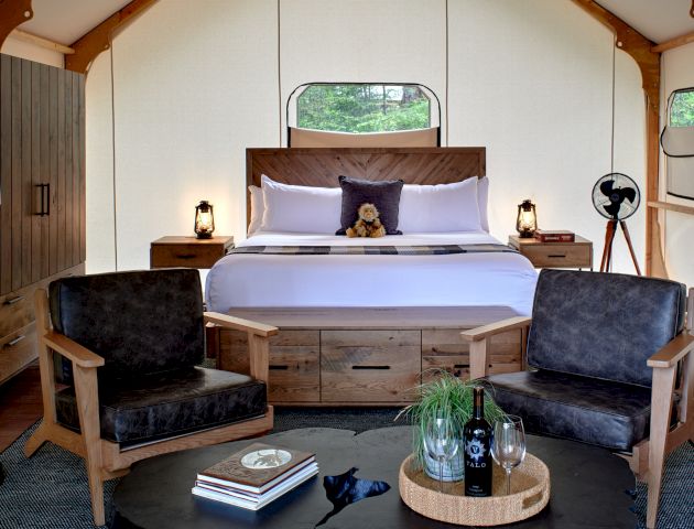 A cozy bedroom with wooden furniture, a double bed, two chairs, decorative items, and a bottle of wine on a table.