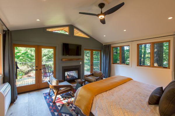 A cozy bedroom with a bed, seating area, fireplace, ceiling fan, and large windows overlooking a wooded area.