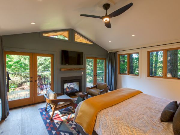 A cozy bedroom with a bed, seating area, fireplace, ceiling fan, and large windows overlooking a wooded area.