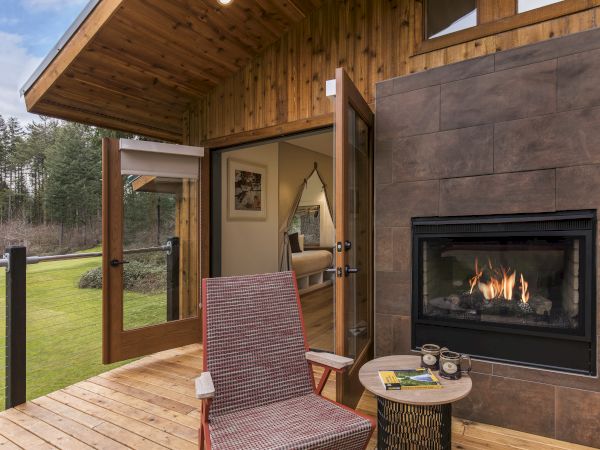 A cozy patio with a wicker chair and small table sits by a modern fireplace, overlooking a green yard, adjoining an inviting interior room.