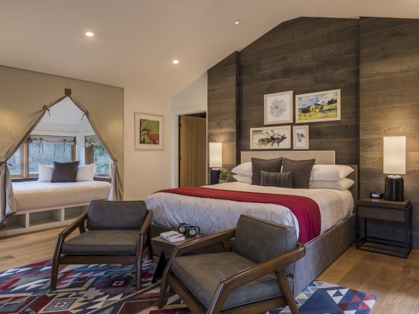 A cozy bedroom with a large bed, red throw, chairs, a decorative rug, and artwork on wood-paneled walls. Window seating is visible.