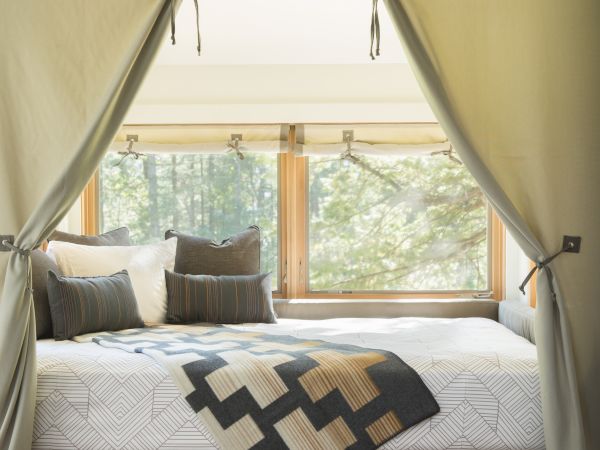 A cozy bed nook with curtains, geometric blanket, pillows, and built-in storage underneath, set against a window with a forest view.