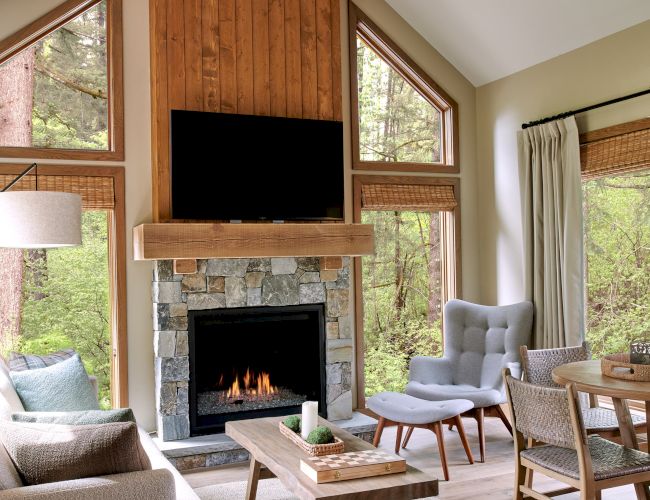 A cozy living room with a tall wooden ceiling, fireplace, TV, sofa, coffee table, and large windows offering a forest view.