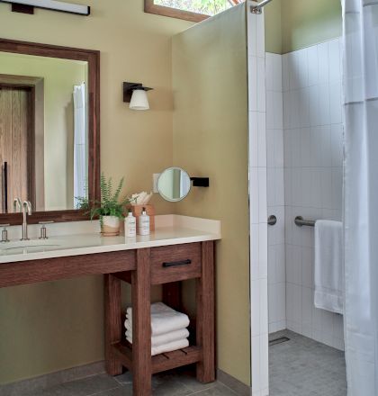 A bathroom with a wooden vanity, mirror, sink, and a shower area. Towels are folded on the shelf below. Natural light is present.