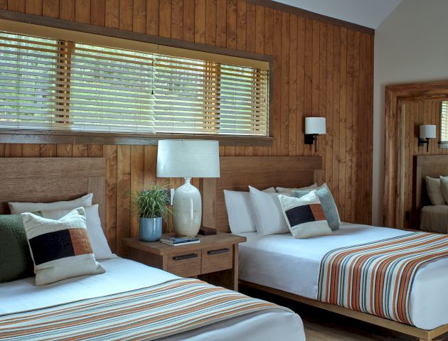 A cozy bedroom with two beds, striped bedding, a lamp, wooden walls, a large mirror, and a window with blinds.