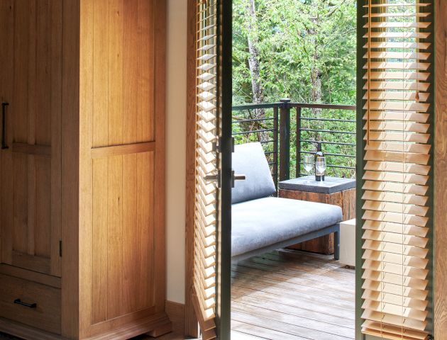 A bedroom opens to a balcony with a wooden door and blinds, featuring outdoor seating and greenery outside.