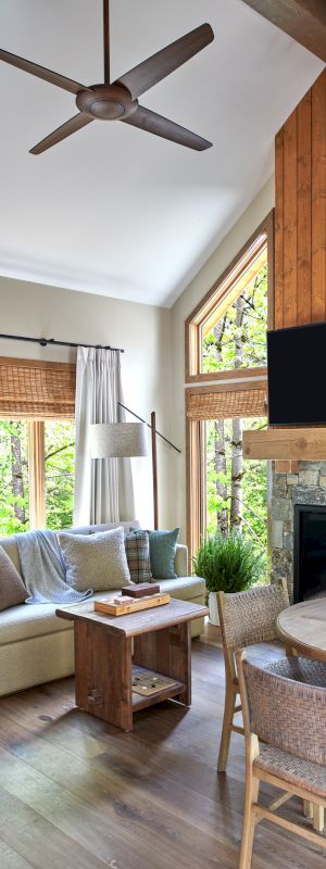 A cozy living room with a sofa, round dining table, fireplace, TV, and ceiling fan. Large windows bring in natural light.