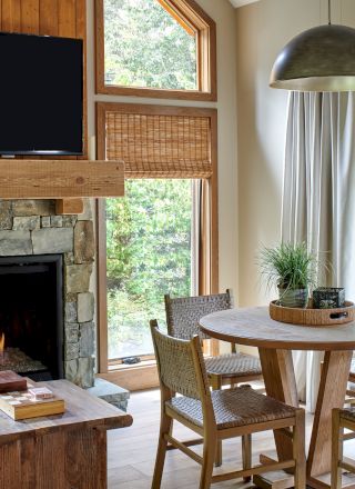 A cozy living area with a stone fireplace, TV, round dining table, four chairs, wood accents, large windows, and nature view.