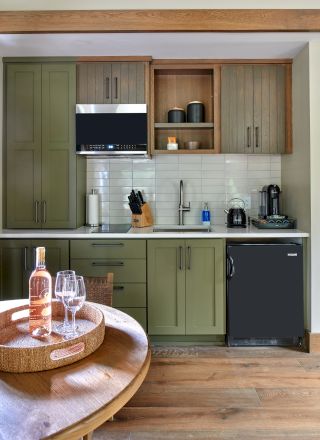 A small kitchen with olive green cabinets, a black mini-fridge, a countertop with kitchen essentials, and a table with wine and glasses.