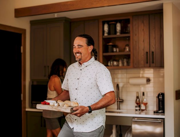 A person is holding a tray in a kitchen with another person nearby. There are beverages on the counter and cabinets in the background.