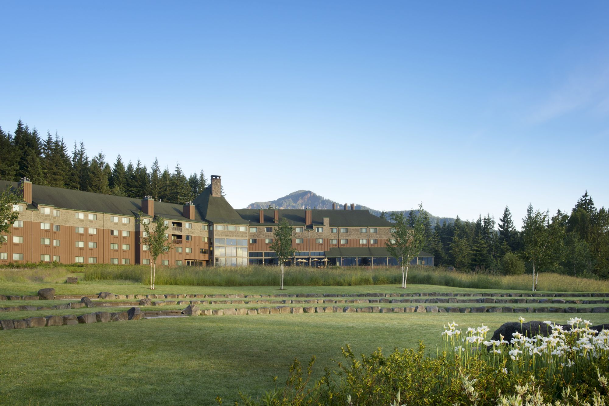 A large building with a sloped roof sits in front of a forest and mountain, surrounded by a grassy area with flowers and trees.
