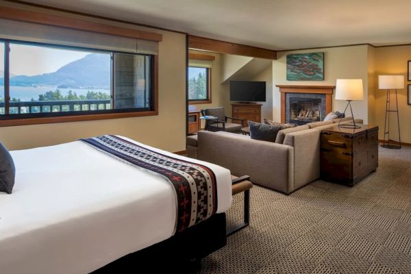 A cozy hotel room with a bed, seating area, fireplace, TV, and a window view of mountains and a lake.