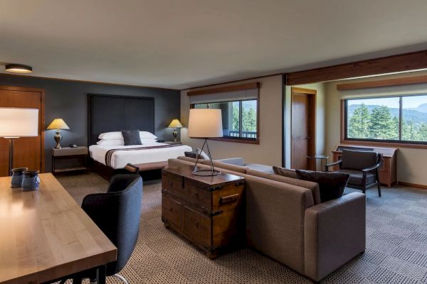 A spacious hotel room with a bed, couch, desk, and large windows offering a view of the greenery outside.