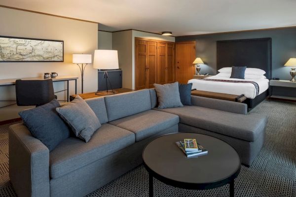 A modern hotel room with a large bed, gray sectional sofa, and a desk with a chair. There are lamps and a coffee table with books.