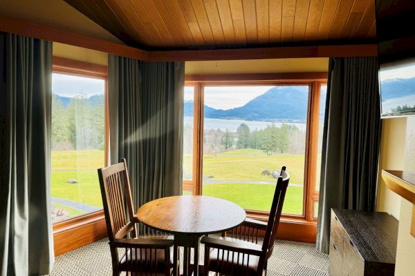 A cozy room with a wooden table and chairs, large windows showcase a scenic mountain and lake view, framed by green trees.