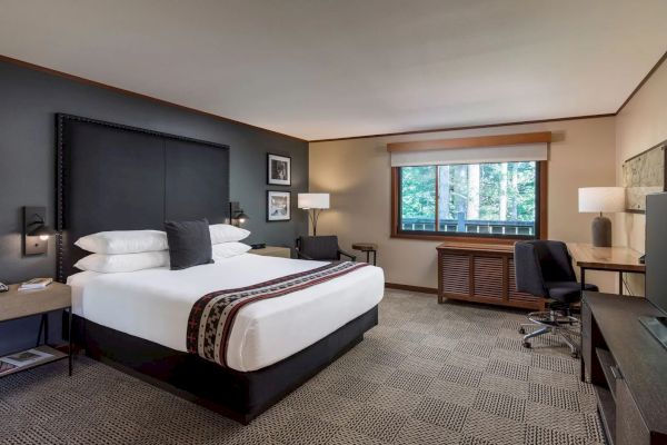 A hotel room with a bed, desk, chair, and window view of trees. There's modern decor, lamps, and framed pictures on the wall.
