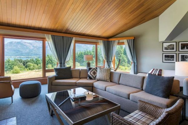 A cozy living room with large windows showing a scenic view, featuring a sectional sofa, glass coffee table, and framed photos on the wall.