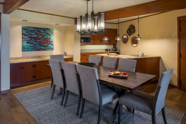 A dining room with a wooden table, gray chairs, a kitchen area, pendant lights, and a colorful fish artwork on the wall ends the scene.