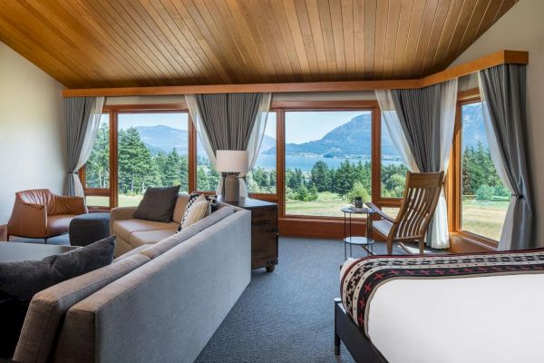 A cozy room with modern furniture, large windows showcasing a scenic view of trees, a lake, and mountains, under a wooden ceiling.