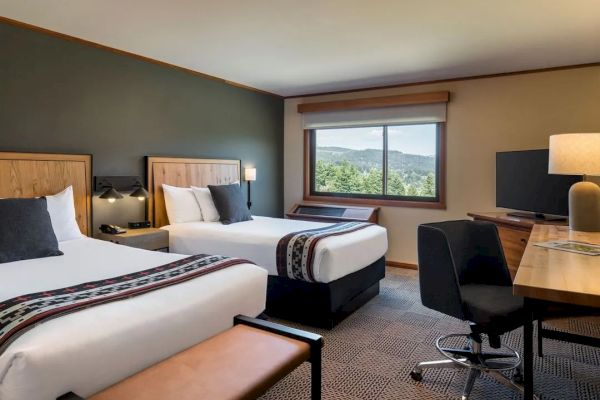 A hotel room with two beds, a desk, a TV, and a window showing a scenic view of trees and mountains.