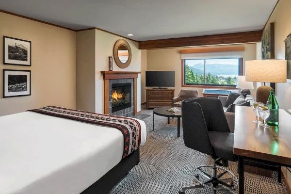 A cozy hotel room features a bed, fireplace, desk, and window view of trees and water, with modern furnishings and decor.
