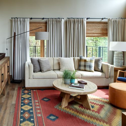 A cozy living room with a sofa, patterned rug, round table, and large windows with curtains and blinds.