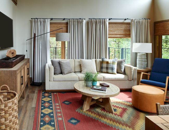 A cozy living room with a sofa, patterned rug, round table, and large windows with curtains and blinds.