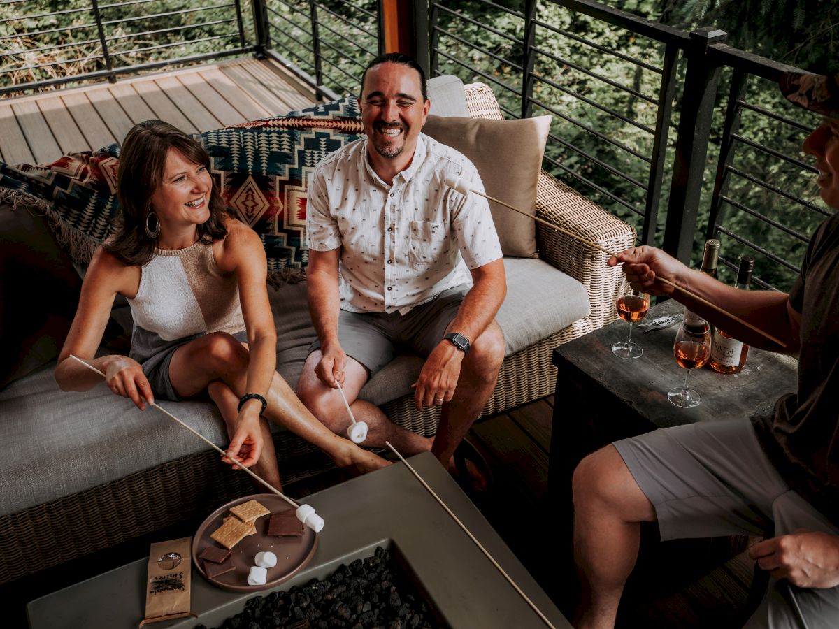 Three people are sitting on a patio sofa, smiling, holding skewers with marshmallows, next to a table with wine and s'mores ingredients.
