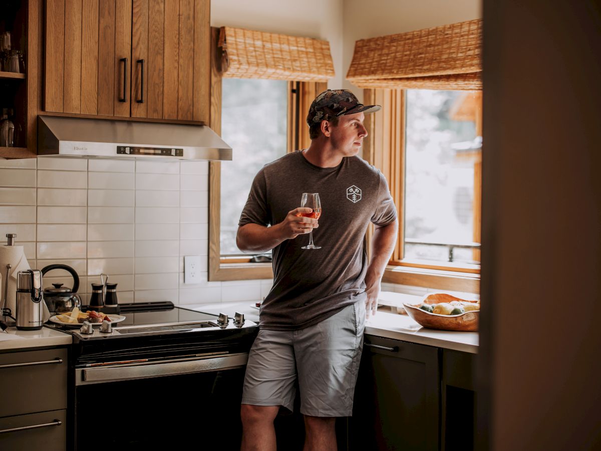 A person stands in a kitchen holding a drink, gazing out of the window, with food items on the counter.
