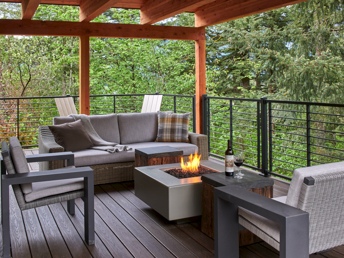 A cozy outdoor patio with modern furniture, a fire pit, and a wine bottle, surrounded by lush greenery and trees, creating a serene atmosphere.