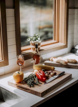 Fresh vegetables, herbs, bread, and a knife on a kitchen counter with a window view and natural light streaming in.